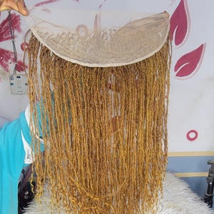 May include: A long, golden-brown wig with a lace front. The wig features numerous, thin dreadlock-style strands. The lace front is a light beige color. The wig is displayed against a white and pink background.
