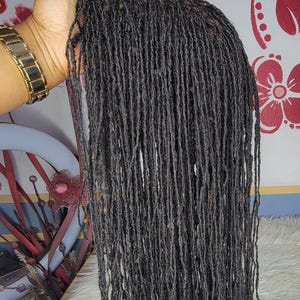 以下が含まれることがあります： 長く細いドレッドヘアのダークグレーのウィッグ。ウィッグは金の時計をした手で持たれています。背景は赤い花の模様の壁です。
