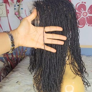 May include: A black micro loc wig on a mannequin head. The wig is styled with small, individual locs. The mannequin head is a light tan color and sits on a white, fluffy surface. A hand is holding a strand of the locs.
