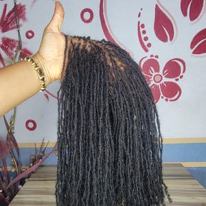 Könnte beinhalten: Eine dunkelgraue Perücke mit zahlreichen kleinen, einzelnen Dreadlocks. Die Perücke wird von einer Hand mit einem goldenen Armband gehalten. Der Hintergrund zeigt eine Wand mit floralen Designs und eine Holzoberfläche.
