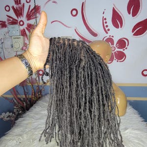 Könnte beinhalten: Ein Bündel grauer Dreadlocks, gehalten vor einer weißen, flauschigen Oberfläche. Die Dreadlocks sind lang und haben ein natürliches, strukturiertes Aussehen. Der Hintergrund umfasst einen Schaufensterpuppenkopf und eine Wand mit roten Blumendesigns.