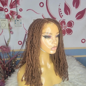 以下が含まれることがあります： マネキンの頭に展示された、肩までの長さのドレッドヘアの茶色のウィッグ。ウィッグは自然なヘアラインで、個々のドレッドヘアでスタイリングされています。マネキンは、花柄と円形のデザインの背景に配置されています。