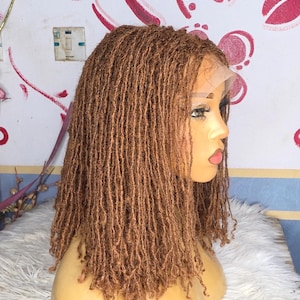 以下が含まれることがあります： レースフロント付きの茶色のウィッグで、多数の小さなドレッドヘアでスタイリングされています。ウィッグは、明るい肌色のマネキンの頭に表示されています。背景は、赤とピンクの装飾要素のある白い壁です。