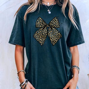 Puede incluir: Camiseta gris oscuro con un gran lazo con estampado de leopardo. El lazo tiene un patrón dorado y negro con detalles en azul. La palabra "Biscuits" está escrita en cursiva encima del lazo. La camiseta es de corte holgado.