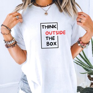 Puede incluir: Camiseta blanca con el texto "THINK OUTSIDE THE BOX" en un rectángulo negro. La palabra "OUTSIDE" es roja. Una prenda casual.