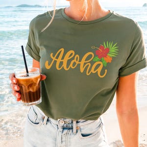 May include: Olive green t-shirt with the word "Aloha" in gold script, and a floral design with red and orange flowers and green leaves. The shirt is worn with light blue denim shorts. A glass of iced coffee is held in the hand.