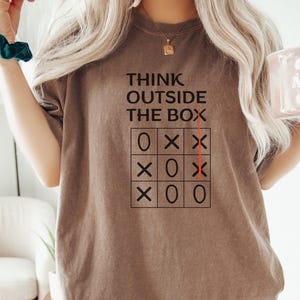 Puede incluir: Camiseta marrón con el texto "THINK OUTSIDE THE BOX" sobre una cuadrícula de tres en raya. La cuadrícula muestra un juego en curso con la "X" ganadora. La camiseta es de color sólido y tiene mangas cortas.