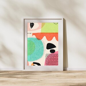May include: Abstract art print in a white frame. The artwork features a colorful design with shapes in pink, green, orange, black, and teal. The print is displayed on a wooden floor against a white wall.