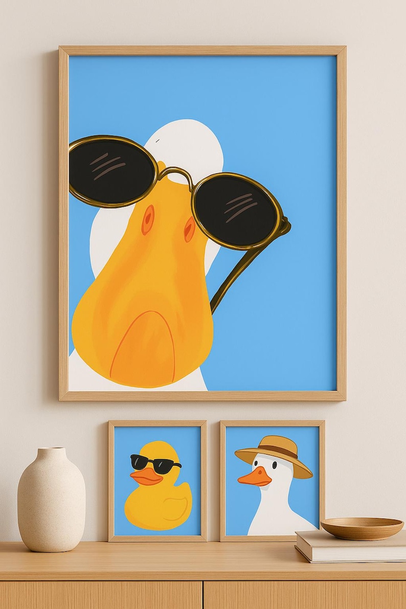 May include: A framed print featuring a cartoon duck wearing sunglasses against a blue background. Below, two smaller framed prints depict a rubber duck with sunglasses and a duck wearing a hat. The artwork is displayed on a wooden surface.
