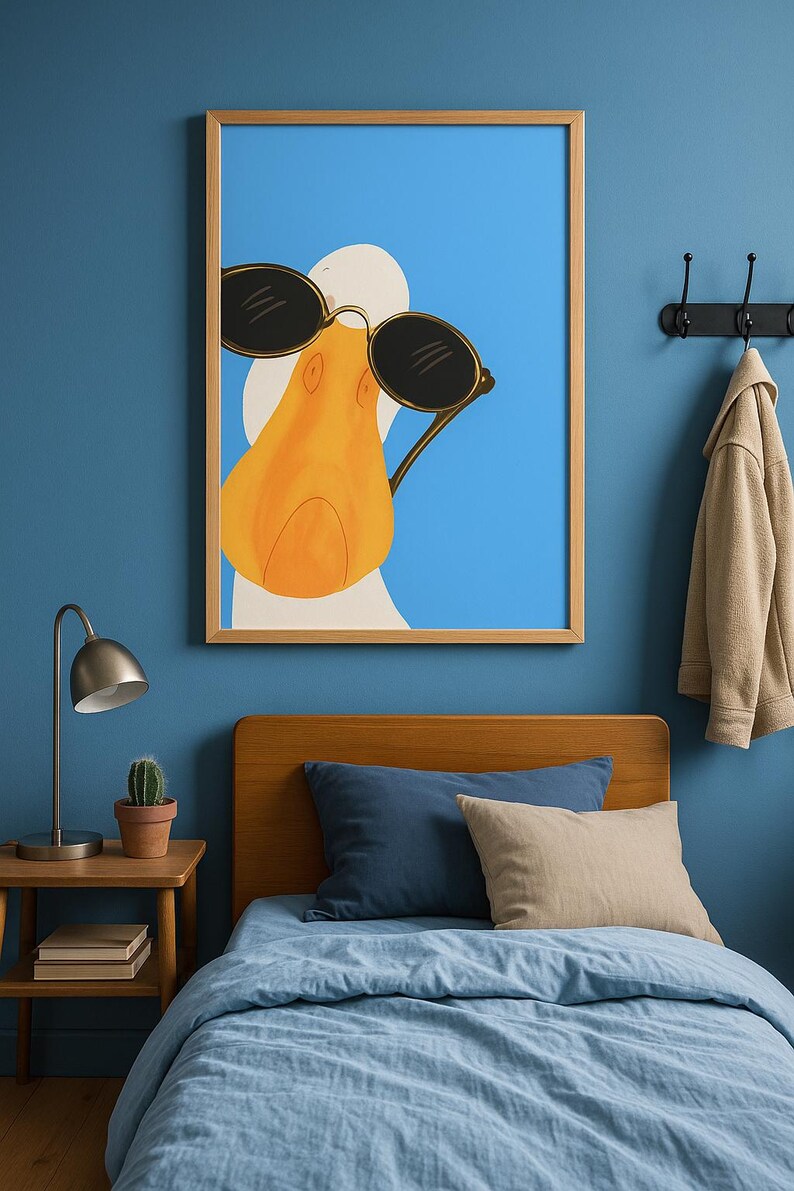 May include: A framed art print featuring a cartoon duck wearing sunglasses against a blue background. The duck has an orange beak and is set in a light wood frame. The image is hanging above a bed with blue bedding and pillows.
