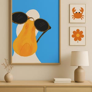 May include: A framed print of a duck wearing sunglasses against a blue background. Two smaller framed prints of a crab and a flower are displayed nearby. The artwork is displayed above a wooden dresser.