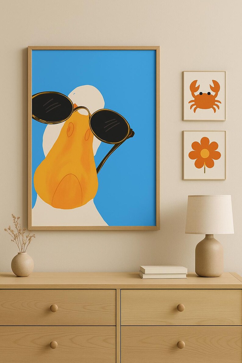 May include: A framed print of a duck wearing sunglasses against a blue background. Two smaller framed prints of a crab and a flower are displayed nearby. The artwork is displayed above a wooden dresser.