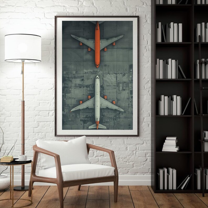 Puede incluir: Una impresi&oacute;n art&iacute;stica a&eacute;rea con dos aviones, uno naranja y otro blanco, sobre un asfalto gris. Los aviones est&aacute;n colocados uno frente al otro. La obra de arte enmarcada se exhibe en una pared de ladrillos blancos.
