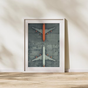 Puede incluir: Una impresi&oacute;n enmarcada que presenta una vista a&eacute;rea de dos aviones en una pista gris. Un avi&oacute;n es naranja, el otro es blanco. La imagen est&aacute; en un marco blanco, sobre una pared blanca y un suelo de madera.