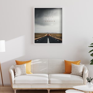 K&ouml;nnte beinhalten: Gerahmter Kunstdruck mit einer Stra&szlig;e unter einem st&uuml;rmischen Himmel und dem Text &bdquo;Start Strong Finish Stronger&ldquo;. Das Bild befindet sich &uuml;ber einem wei&szlig;en Sofa mit orangefarbenen und grauen Kissen. Das Sofa befindet sich in einem Raum.