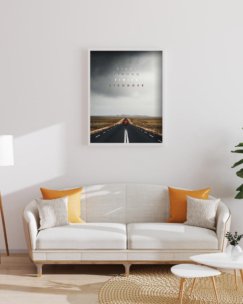 K&ouml;nnte beinhalten: Gerahmter Kunstdruck mit einer Stra&szlig;e unter einem st&uuml;rmischen Himmel und dem Text &bdquo;Start Strong Finish Stronger&ldquo;. Das Bild befindet sich &uuml;ber einem wei&szlig;en Sofa mit orangefarbenen und grauen Kissen. Das Sofa befindet sich in einem Raum.