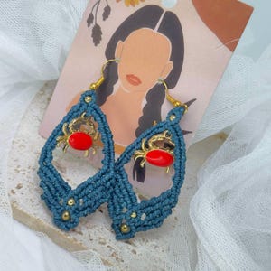 May include: Teal macrame earrings with gold-colored crab charms featuring red enamel details. The earrings have gold-colored hooks and small gold beads. The earrings are displayed on a decorative card with an illustration of a woman.