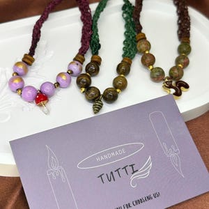 Puede incluir: Collares Tutti hechos a mano con varios diseños de cuentas. Un collar presenta cuentas moradas con un dije de hongo rojo. Otro tiene cuentas verdes con un dije de piña. El tercer collar tiene cuentas marrones con un dije de ardilla. Una tarjeta de visita dice "Handmade Tutti, ¡Gracias por elegirnos!"