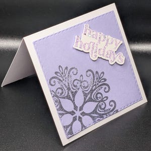 May include: A handmade greeting card with a lavender background and a decorative snowflake design. The card features the text "happy holidays" in glittery letters. The card is folded and has a white border.