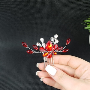 May include: A silver hair comb adorned with vibrant red and clear crystal-like embellishments. The design features a central cluster of large, faceted red stones surrounded by smaller red and clear accents, creating a sparkling, floral-inspired accessory.