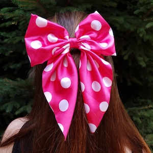May include: A large, bright pink hair bow with white polka dots. The bow is made of a shiny fabric and is tied in the center. The bow is worn in the hair, with the wearer's brown hair visible.