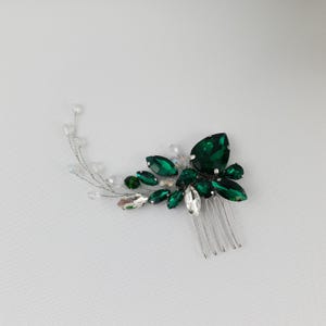 May include: A hair comb featuring a cluster of emerald green and clear crystal-like stones. The stones are arranged in a floral design, with a silver comb base. The comb is designed for formal occasions.
