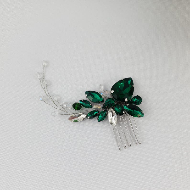 May include: A hair comb featuring a cluster of emerald green and clear crystal-like stones. The stones are arranged in a floral design, with a silver comb base. The comb is designed for formal occasions.