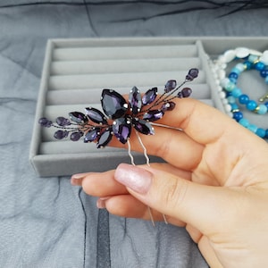 Può includere: Un fermaglio per capelli viola scuro con un design floreale. Il fermaglio presenta grandi cristalli a forma di goccia e cristalli più piccoli e rotondi. I cristalli sono incastonati su una base di filo scuro con due forcine d'argento.