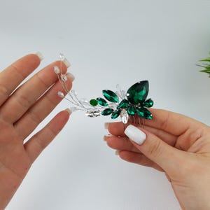 May include: A hair comb featuring a cluster of emerald green and clear crystal rhinestones. The comb has a silver wire design with small clear beads and is designed for formal occasions.