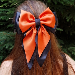 Pode incluir: Um grande laço de cabelo de duas cores. O laço apresenta um centro laranja vibrante com bordas e caudas pretas. O laço é feito de um material brilhante, semelhante ao cetim. O laço é usado no cabelo.