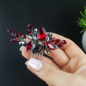 Puede incluir: Un peine decorativo para el cabello con cristales rojos, negros y plateados. Los cristales están dispuestos en un diseño floral y sujetos a un peine plateado. El peine se sostiene en una mano sobre un fondo oscuro.