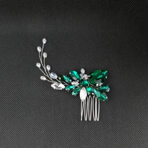May include: A silver hair comb with a floral design. The comb features a cluster of emerald green and clear crystal-like stones. The stones are arranged to resemble leaves and flowers, with a delicate silver wire stem.