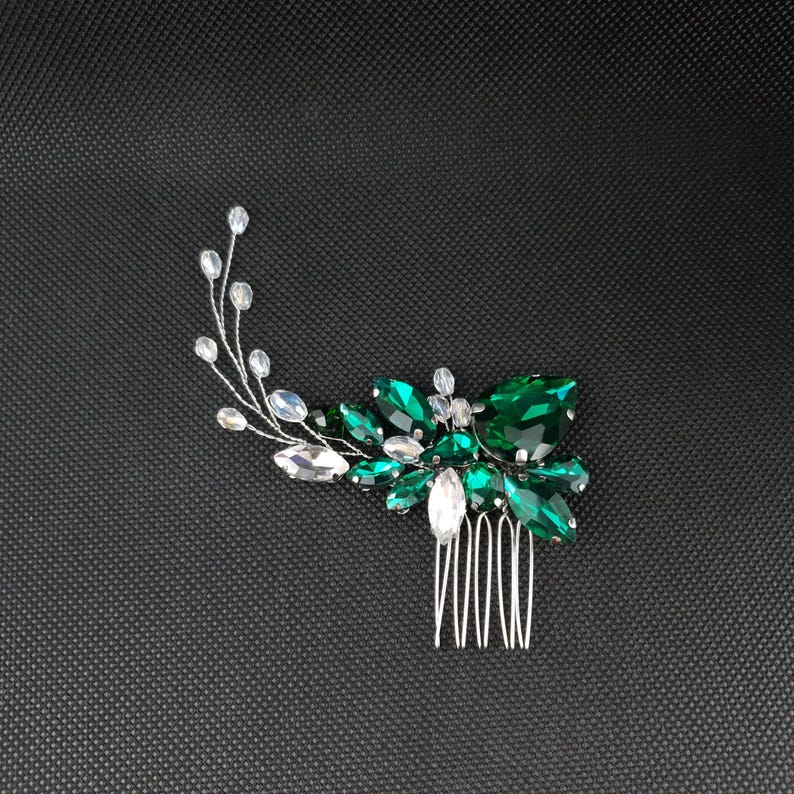May include: A silver hair comb with a floral design. The comb features a cluster of emerald green and clear crystal-like stones. The stones are arranged to resemble leaves and flowers, with a delicate silver wire stem.