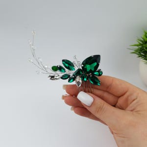 May include: A hair comb featuring a cluster of emerald green and clear crystal-like stones. The comb has a silver-colored metal base with a series of teeth for securing it in the hair. The crystals are arranged in a floral or leaf-like pattern.