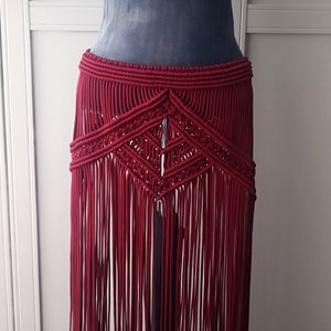 Puede incluir: Una falda larga de macramé de color burdeos con un patrón geométrico y flecos. La falda está hecha de cordón grueso y tiene un estilo bohemio.