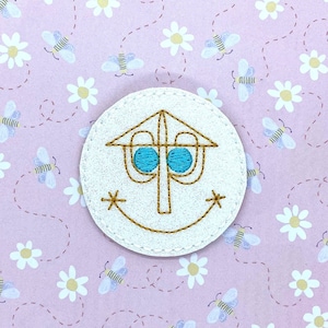 May include: A white, circular embroidered patch featuring a stylized face with blue eyes and a smiling mouth. The patch has a brown outline and is set against a pink background with a pattern of bees and flowers.