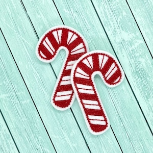 May include: Two red and white candy cane shaped felt ornaments on a light blue wooden background.