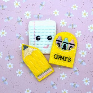 May include: Three felt appliques in the shape of school supplies: a yellow pencil, a white sheet of paper with lines, and a yellow box of crayons. The crayons are visible inside the box. The appliques are on a pink background with a pattern of white daisies and bees.