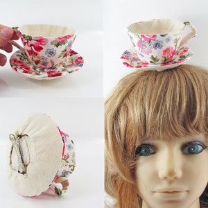 May include: A white teacup and saucer hair clip with a floral pattern in pink, red, and blue. The clip is made of fabric and has a metal clasp on the back.