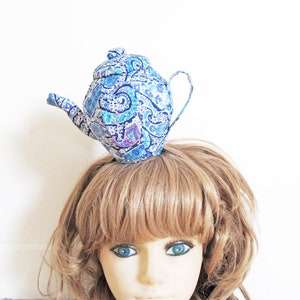 Può includere: Un cappello stravagante a forma di teiera blu paisley. Il cappello presenta un beccuccio curvo, un manico e un coperchio ed è decorato con un motivo ripetuto di disegni paisley blu, bianchi e viola. Il cappello è progettato per essere indossato sulla testa.