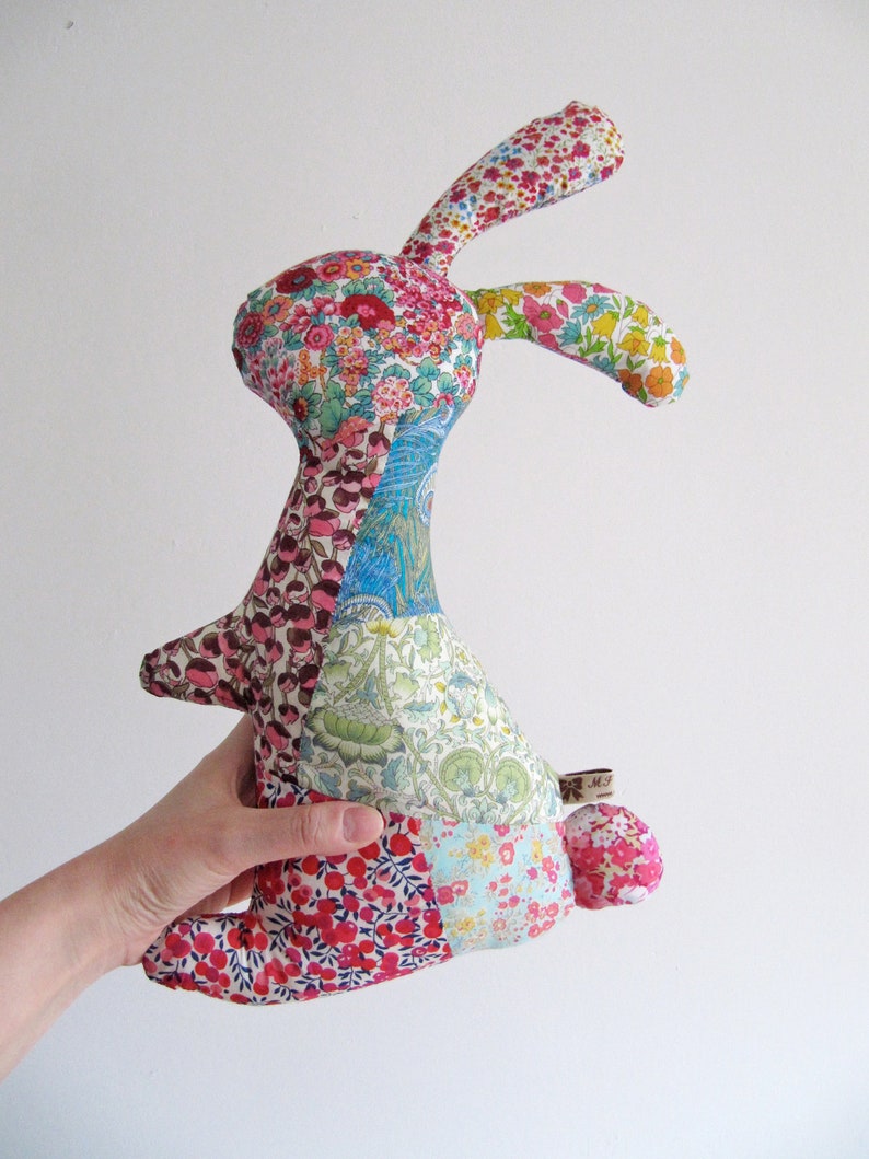 May include: A patchwork fabric bunny toy with a floral pattern. The bunny is made of different coloured fabrics, including pink, blue, green, and red. The bunny has a long, floppy ear and a small tail.