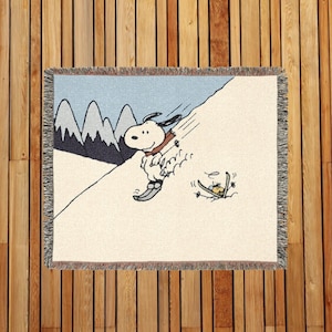 Puede incluir: Una manta tejida con una ilustración de dibujos animados de Snoopy esquiando. El diseño muestra a Snoopy con esquís, una pendiente nevada, un cielo azul claro y montañas. La manta tiene un borde con flecos.