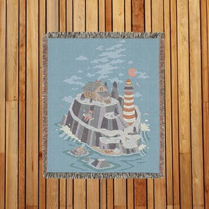 May include: A woven throw blanket featuring an island scene with a lighthouse, house, and skull-shaped rock formation. The blanket has a light blue background with a fringed edge. The artwork depicts a sunset and ocean waves.