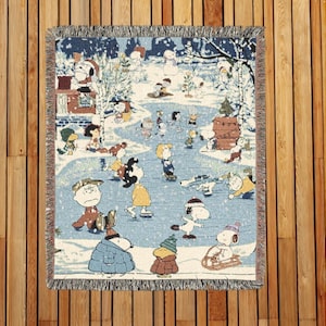 Puede incluir: Una manta tejida con una escena invernal con personajes de dibujos animados patinando y jugando en la nieve. La manta tiene un fondo azul con un borde de flecos y se muestra sobre una superficie de madera.