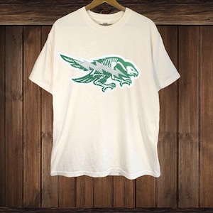 May include: Cream-colored t-shirt featuring a green and white graphic of a bird with a lightning bolt design. The bird's skeletal structure is visible. The shirt is hanging on a wooden hanger against a wooden background.