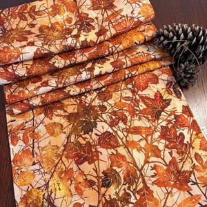 May include: A folded table runner with a fall leaf pattern in shades of orange, brown, and yellow. The fabric features detailed illustrations of maple and oak leaves, creating a seasonal autumn theme. A pine cone rests on the dark wooden surface.