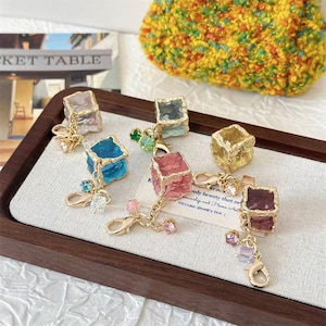 Könnte beinhalten: Eine Sammlung farbenfroher, würfelförmiger Anhänger mit goldfarbener Hardware. Die Anhänger sind in verschiedenen Farben erhältlich, darunter Rosa, Blau und Gold, und sind mit kleinen Kristallen verziert. Die Anhänger werden auf einem Holztablett präsentiert.