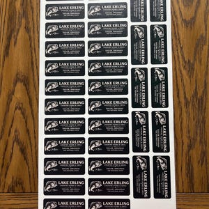 May include: Sheet of black and white rectangular stickers. Each sticker features the text "LAKE ERLING COUNTRY STORE & GRILL TAYLOR, ARKANSAS THE NATURAL STATE" with a fish graphic. The stickers are on a white background.