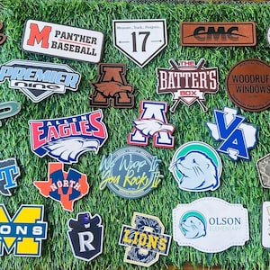 May include: A collection of various patches and emblems, featuring logos and text. Designs include sports team logos, business names, and phrases like "Can't Stop Won't Stop." The patches are in various shapes and colors, displayed on a green surface.