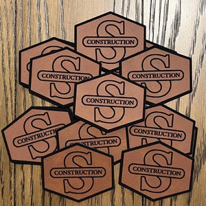 May include: A collection of tan, hexagonal patches featuring a black border and a large black letter S. Each patch displays the word CONSTRUCTION in black capital letters inside a horizontal bar that overlays the center of the letter S.